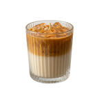 An iced coffee-flavored candle in a clear ribbed glass tumbler, filled with a layered brown and cream liquid and topped with ice cubes, on a solid white background.